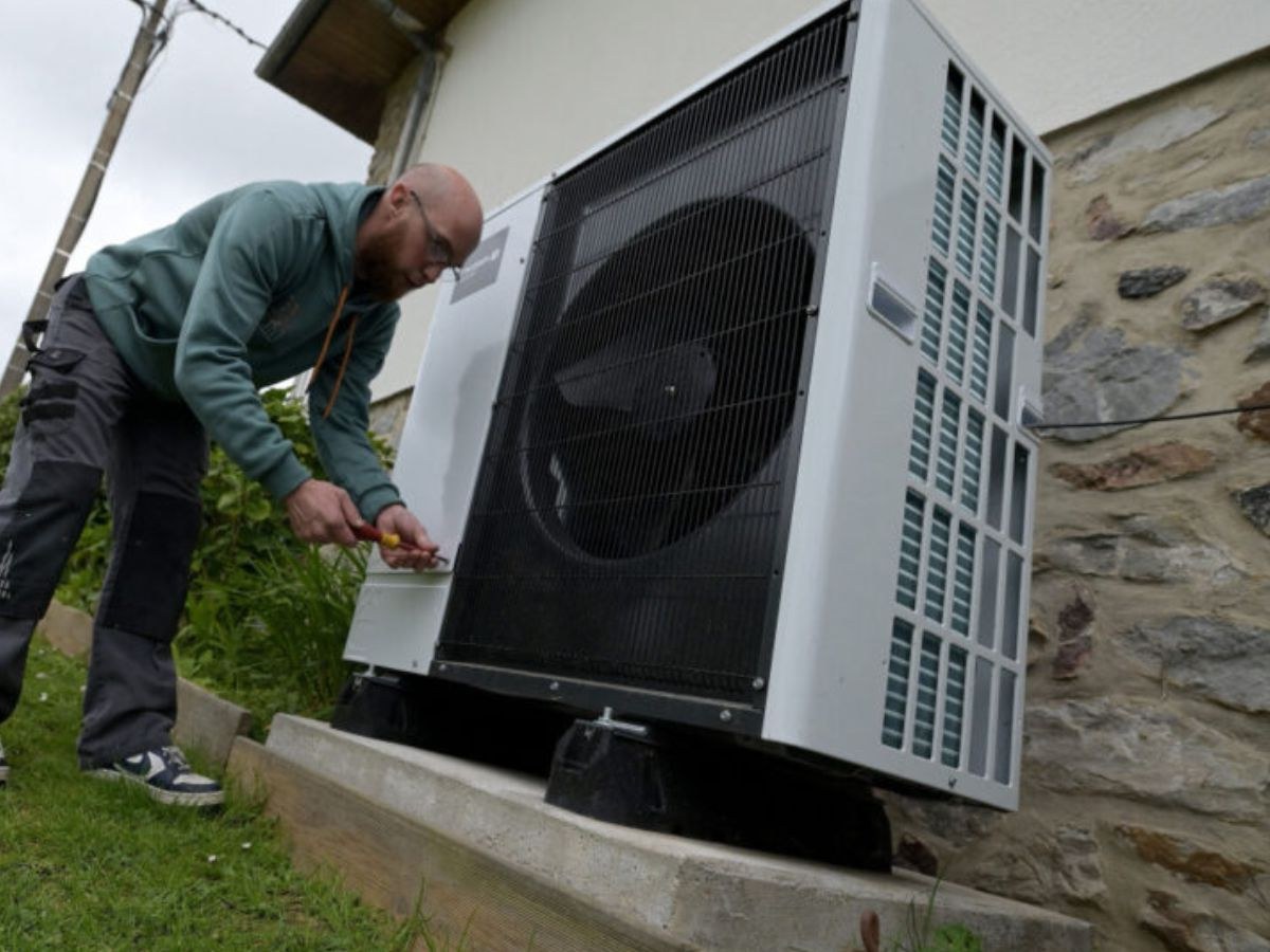 Technicien en sweat vert visse le panneau d'une pompe à chaleur extérieure, grille de ventilateur noire.