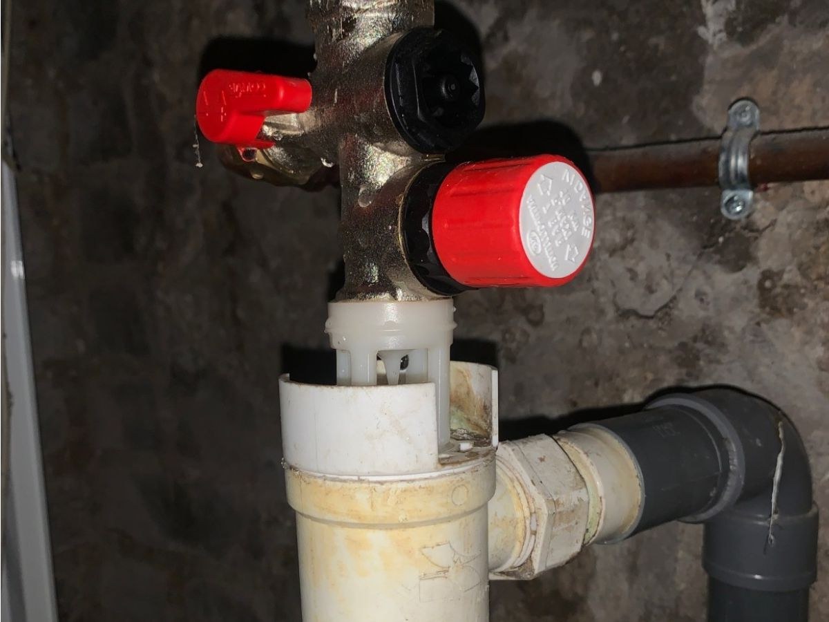Groupe de sécurité en laiton, bouton rouge de vidange, entonnoir blanc et tube PVC contre un mur brut.