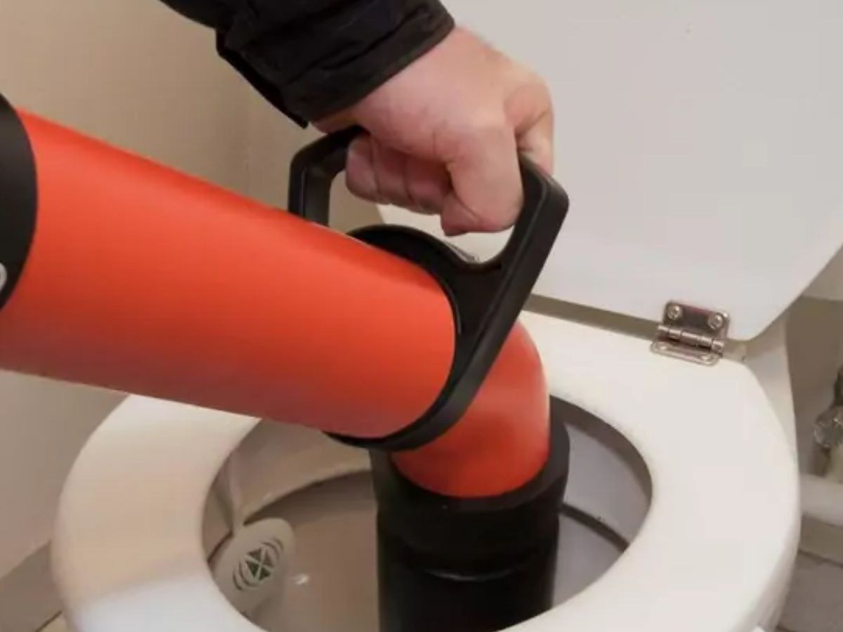 Main gantée enfonçant un déboucheur orange dans la cuvette d'un WC, abattant relevé contre le mur.