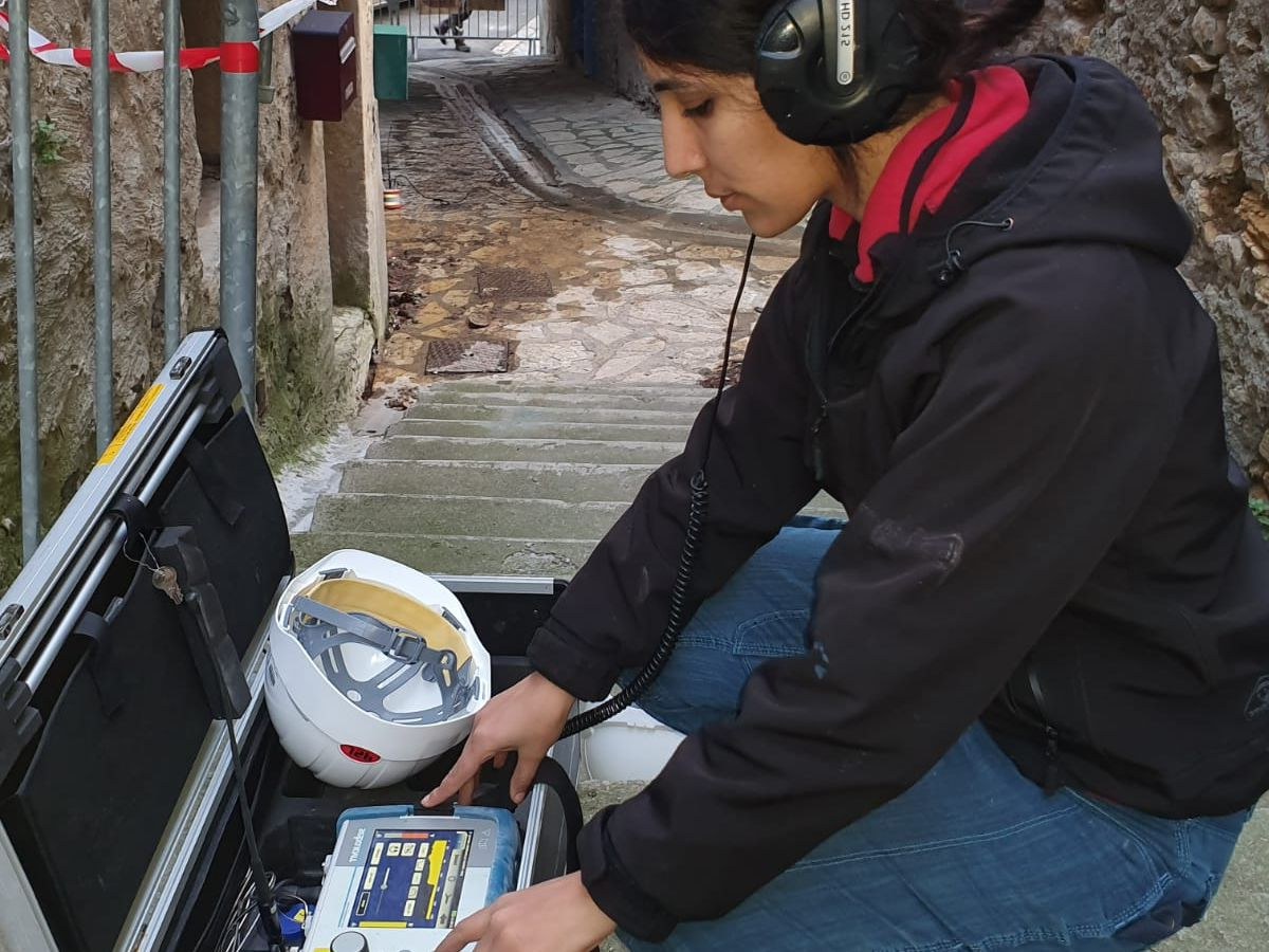 Technicienne à genoux, casque sur les oreilles, devant un boîtier d'écoute dans une ruelle pavée.