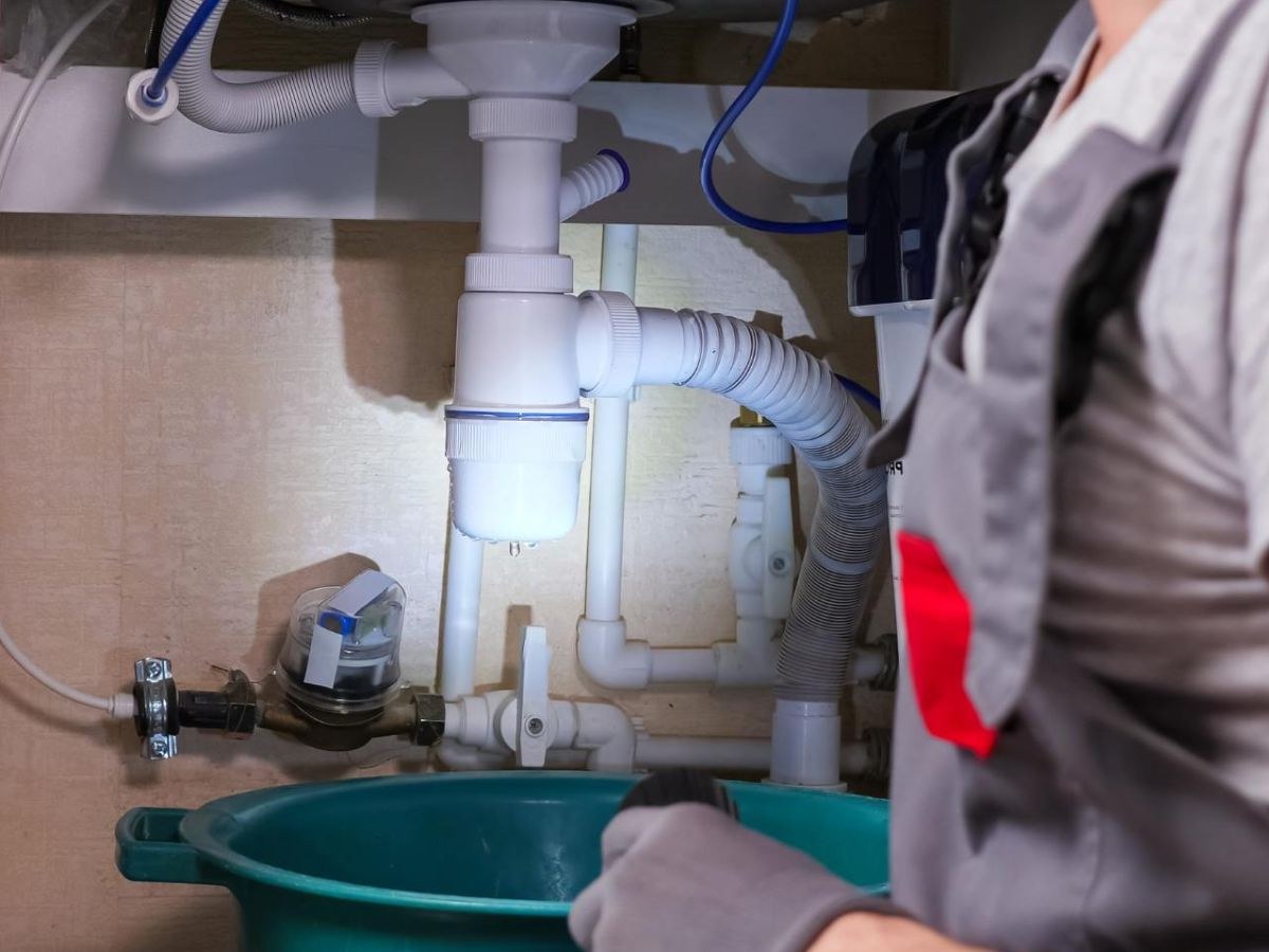 Goutte suspendue sous un siphon blanc, recueillie dans une bassine verte, plombier éclairant le meuble.