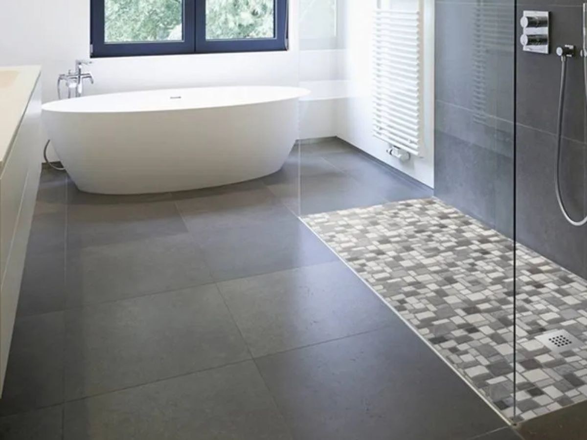 Douche mosaïque gris-blanc à côté d'une baignoire îlot ovale, sol anthracite et fenêtre.