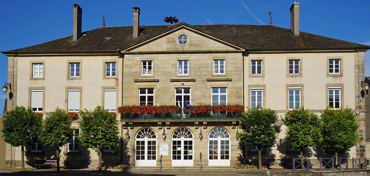 Mairie en pierre claire, balcon fleuri de géraniums, drapeaux français et européen derrière une rangée d'arbres.