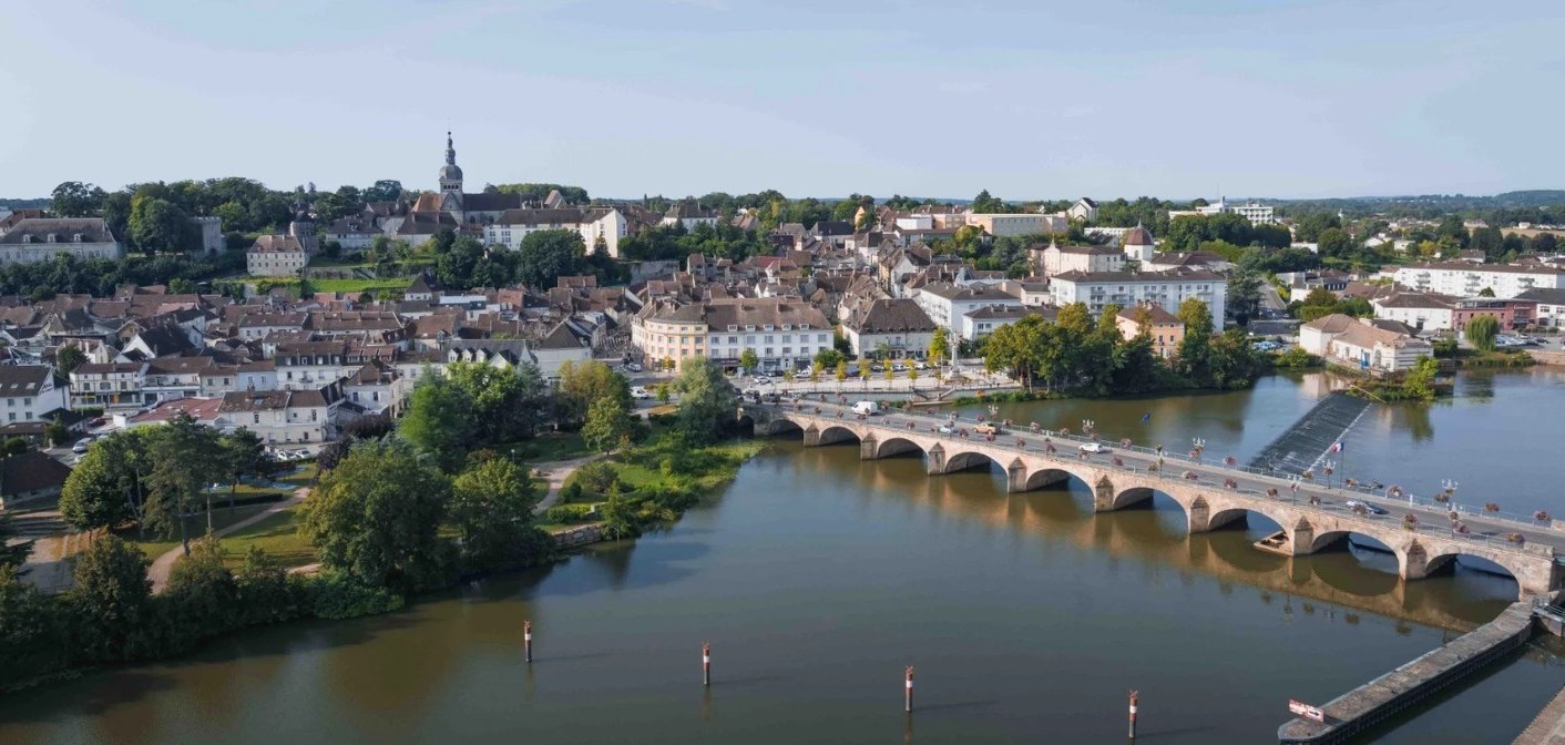Rivière traversée par un pont de pierre à arches, face à des toits rouges et un clocher.
