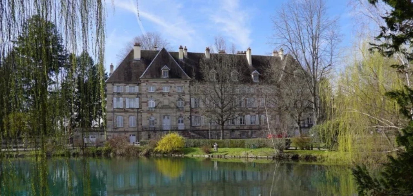 Manoir en pierre aux volets blancs, reflété dans un étang calme, encadré de saules pleureurs.