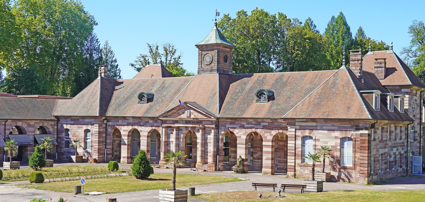 Thermes en grès rose et beige, tour à horloge et parvis planté sous un ciel bleu.