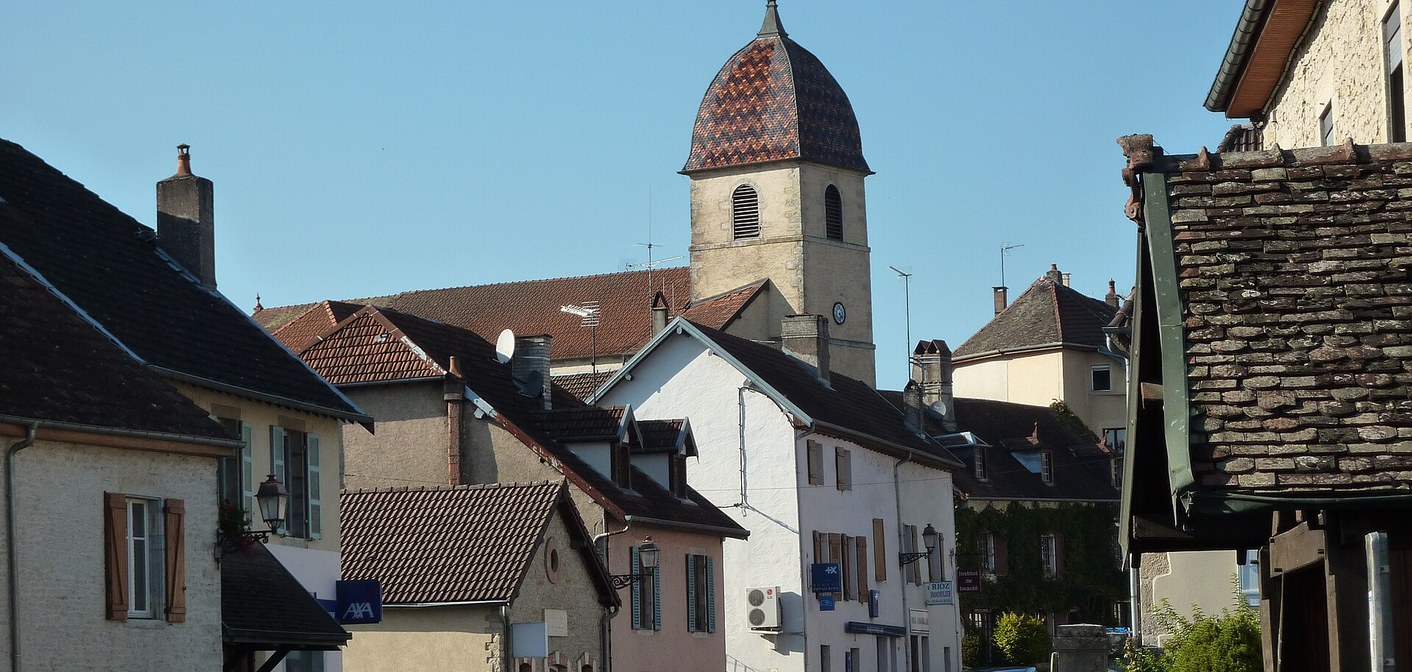 Toits de village et clocher comtois vernissé vus depuis la rue principale sous un ciel bleu.