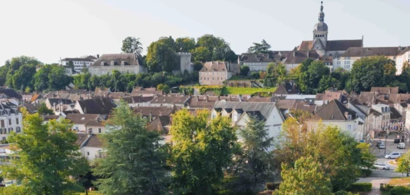 Ville en pente aux toits de tuiles rouges, clocher à bulbe sombre et arbres entre les façades claires.
